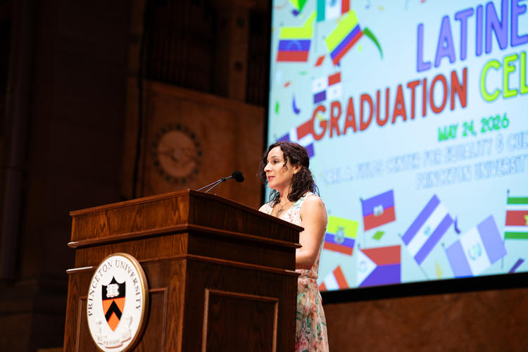 Prof. Anais Holgado Lage wearing a floral dress in front of a podium and with a screen behind her that says Latine Graduation, giving a speech