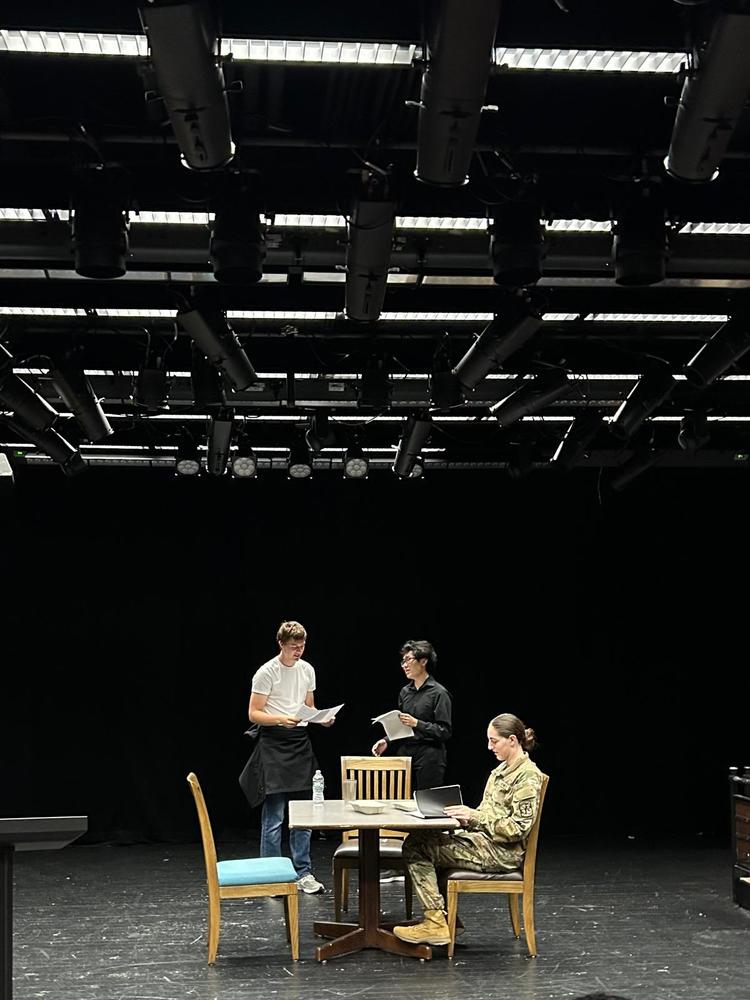 A theatre play is taking place with three students, one sitting at a table and two talking behind, with a black background